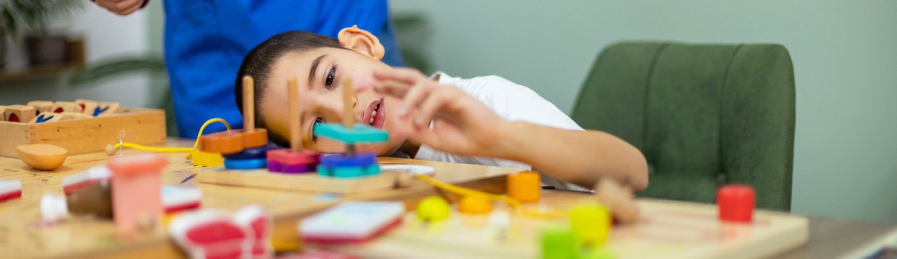 A boy rests his head on the table and reaches for colourful wooden shapes while a therapist stands behind him