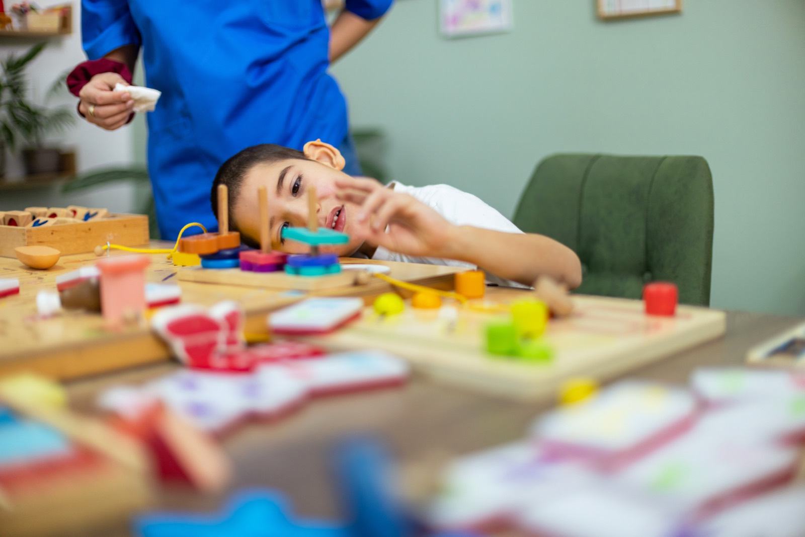 A boy reaches for colourful wooden shapes while a therapist stands behind him