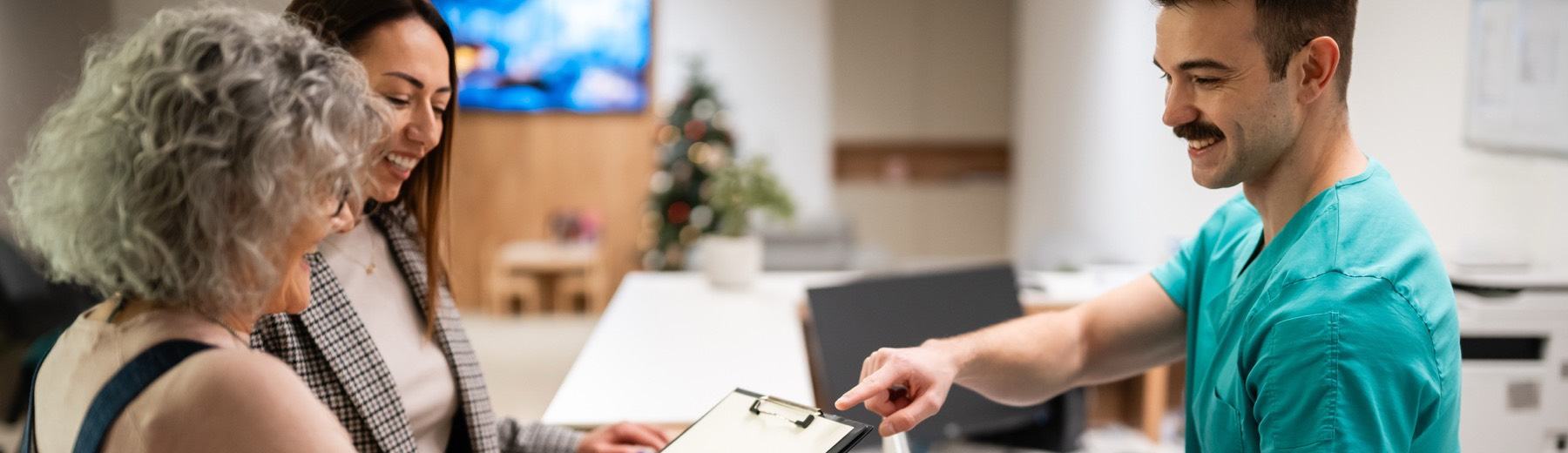 A grandmother and mother fill in a clipboard at a clinic reception desk as a nurse points to the form