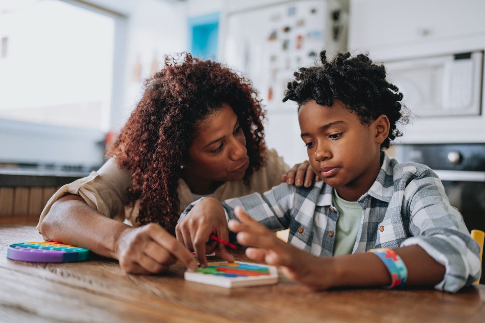 A mother guides her son's hands on a wooden puzzle at the kitchen table