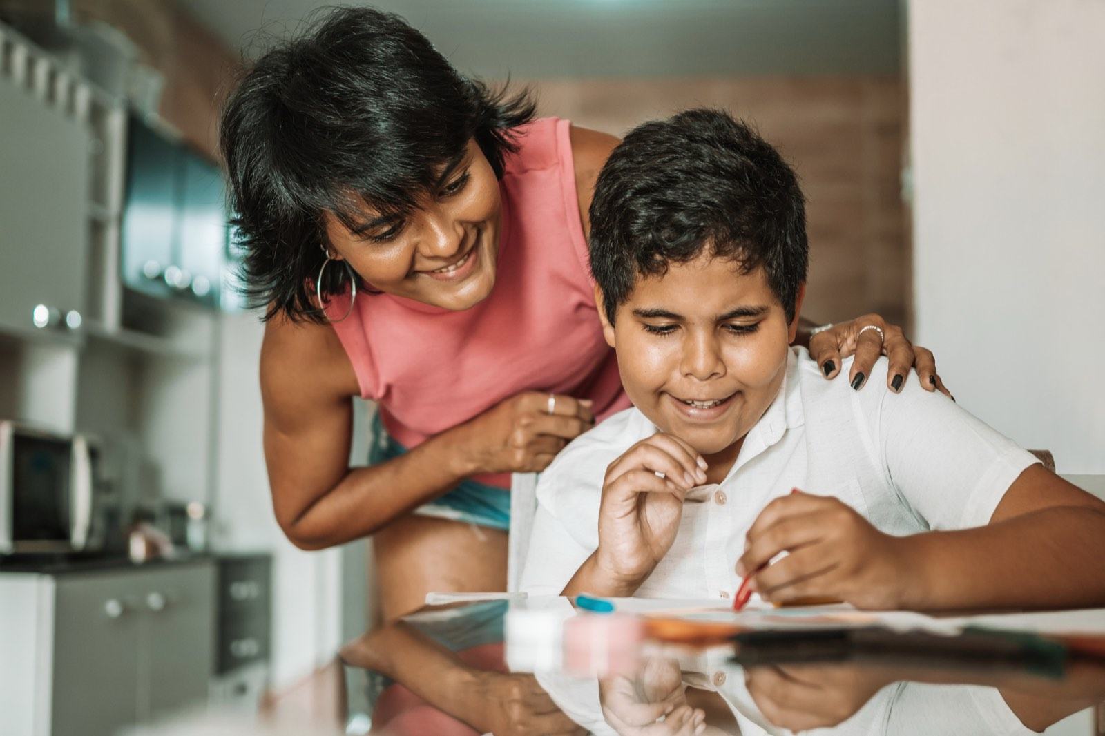 A mother leans over her son's shoulder as he draws at the kitchen table