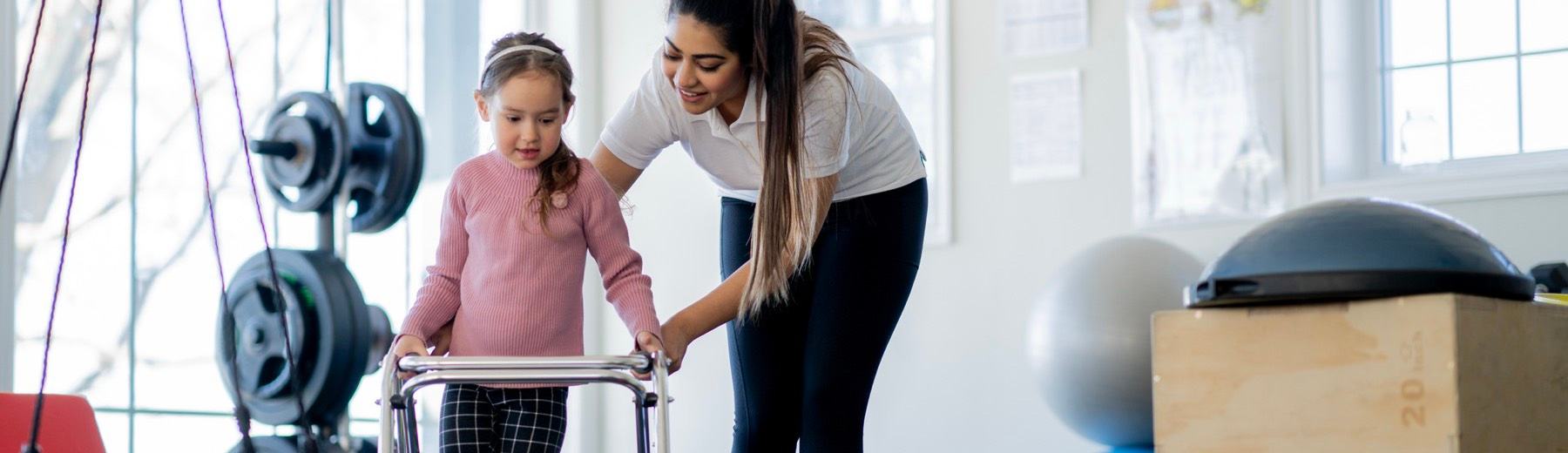 A physiotherapist steadies a young girl using a walker between cones in a therapy gym