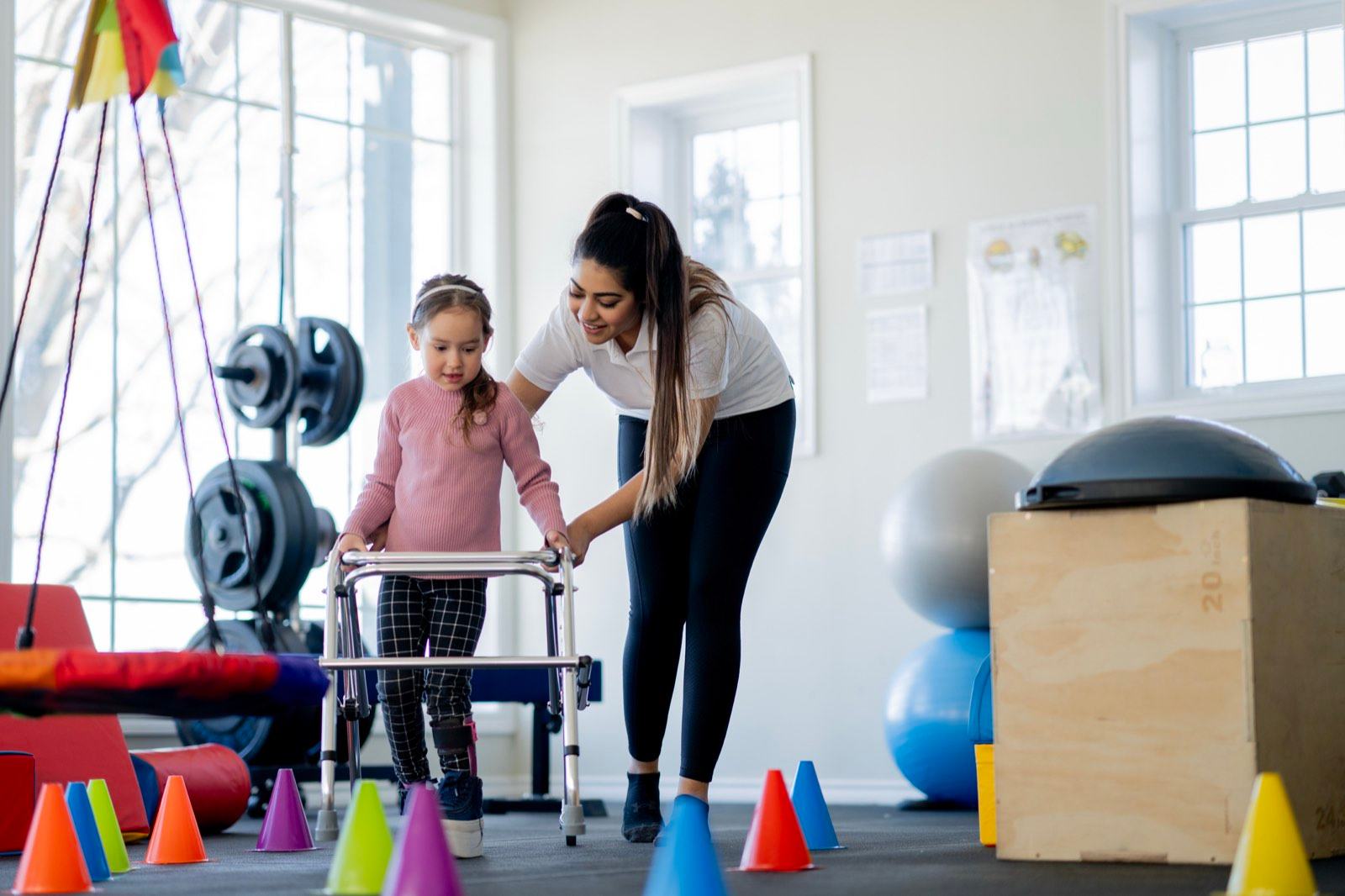 A physiotherapist steadies a young girl using a walker between cones in a therapy gym