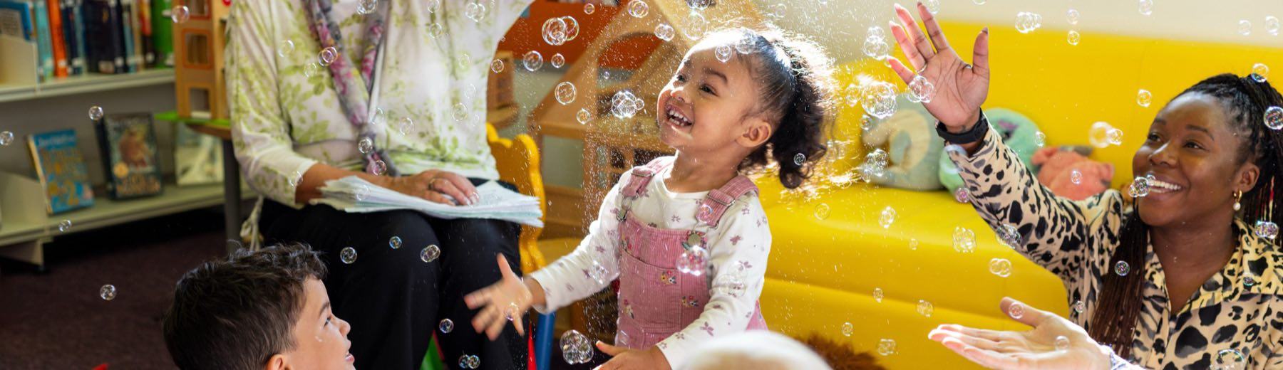 Two carers and three toddlers in a playgroup, laughing as soap bubbles fall around them