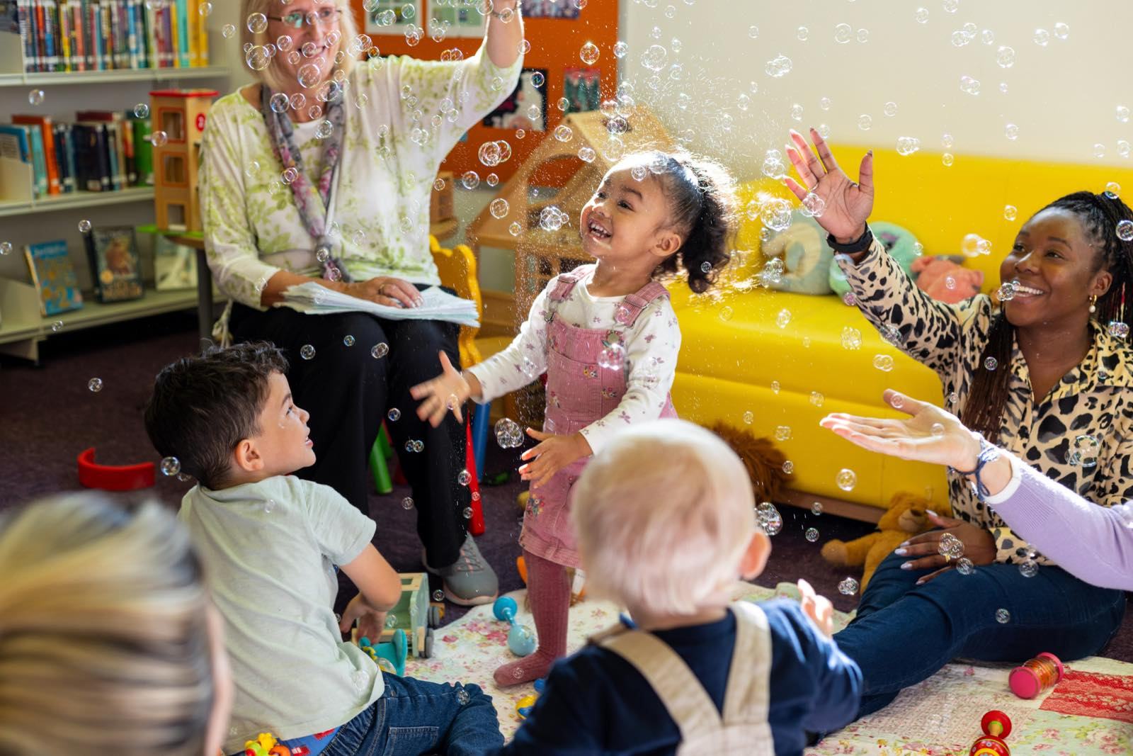 Two carers and three toddlers in a playgroup, laughing as soap bubbles fall around them