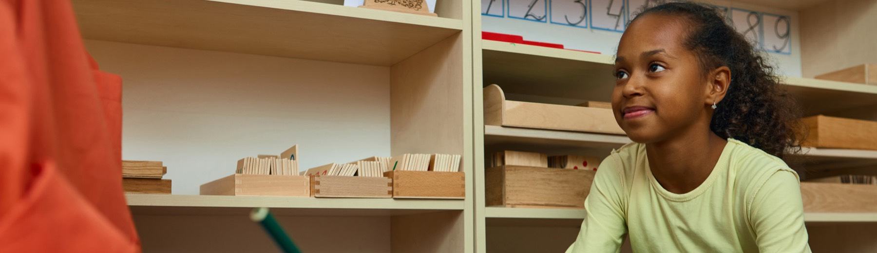 A girl smiles at a teacher taking notes in a classroom lined with wooden learning materials