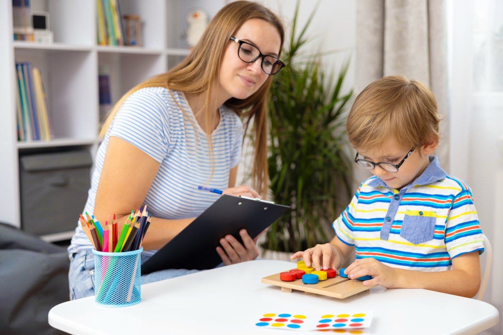 A therapist with a clipboard watches a boy in glasses work through a peg board